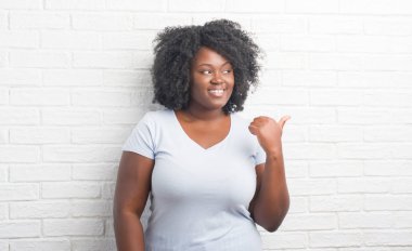Genç Afro-Amerikan artı boyutu kadın arıyorum ve başparmak yanında yukarıyı mutlu yüz ile gülümseyen beyaz tuğla duvarın üzerinden.