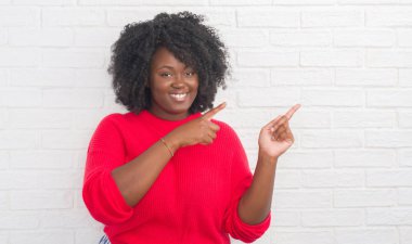 Gülümseyen ve iki elleri ve parmakları ile tarafına işaret kamera bakarak genç Afro-Amerikan artı boyutu kadın beyaz tuğla duvar.