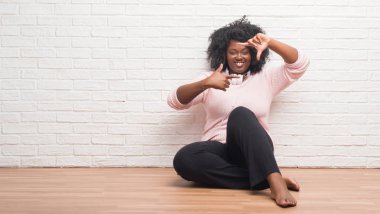 Elle yapma çerçeve ve parmakları ile mutlu yüz gülümseyen kulaklıklar giyen katta oturan genç Afro-Amerikan kadın. Yaratıcılık ve Fotoğrafçılık kavramı.