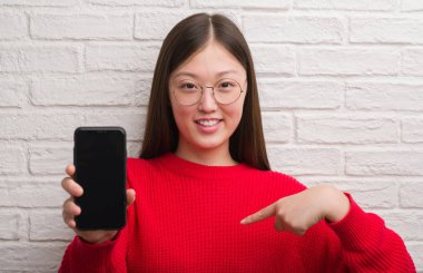 Tuğla duvar gösteren smartphone sürpriz yüz işaret parmağı kendine Genç Çinli kadın