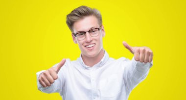 Genç yakışıklı sarışın adam el ile pozitif jest yapıyor onaylama başparmak gülümseyen ve başarı için mutlu. Kamera, kazanan jest arıyorsunuz.