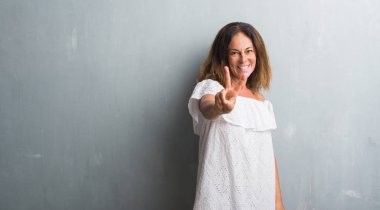 Orta yaş İspanyol zafer işareti yapıyor parmak gösterilen kamera seyir gülümseyen gri grunge duvarın üzerinden duran kadın. İki numaralı.