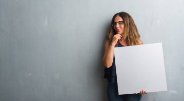 Gri grunge duvar holding boş afiş ciddi bir yüz düşünme hakkında soru, çok karışık bir fikir üzerinde duran orta yaş İspanyol kadın