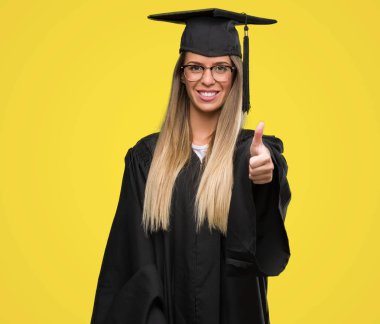 Güzel genç kadın gözlük ve Mezuniyet cübbesi kocaman bir gülümseme ile ok işareti yaparken mutlu parmak ile mükemmel işareti yukarı başparmak