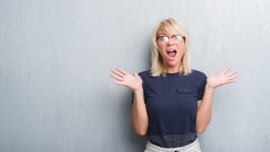 Grunge yetişkin beyaz kadın deli kutluyor gözlük takan duvar gri ve çığlık kaldırdı ve açık gözleri heyecanlı silah ile başarı için hayrete düşürdü. Kazanan kavramı