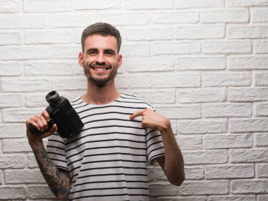 Filme holding video kamera sürpriz yüz işaret parmağı beyaz tuğla duvarın üzerinden kendisine duran genç adam