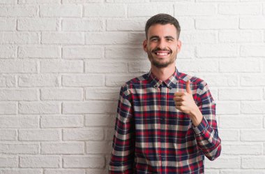 Hareketi el ile mutlu yaşasın yapıyor beyaz tuğla duvarın üzerinden ayakta genç yetişkin adam. Kameraya bakarak başarı gösteren ile ifade onaylama.