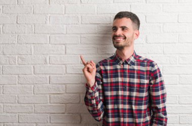 Kameraya bakarak yan el ve parmak ile işaret eden genç yetişkin adam yüzünde kocaman bir gülümseme ile beyaz tuğla duvarın üzerinden ayakta.