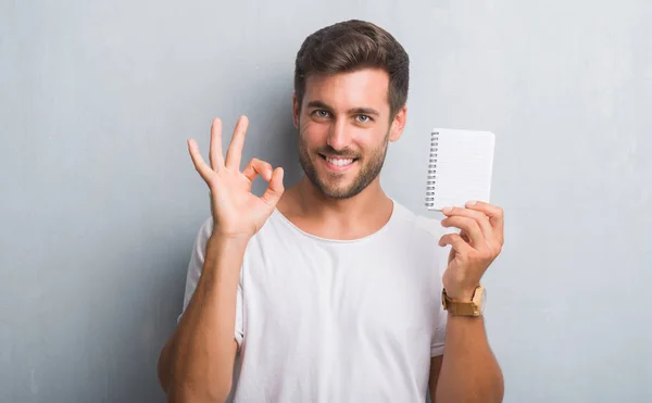 英俊的年轻人在灰色垃圾墙持有空白笔记本做 标志与手指 优秀的符号 — 图库照片