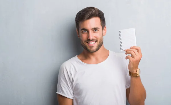英俊的年轻人在灰色垃圾墙持有空白笔记本与一个快乐的脸站在微笑着自信的微笑显示牙齿 — 图库照片