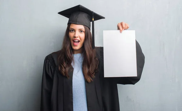 若いブルネットの女性グランジに灰色の驚き顔 恐怖表現と興奮とショックで怖い学位を保持する大学院の制服を着て壁 — ストック写真