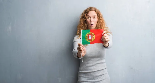 灰色垃圾墙上的年轻红头发的女人拿着葡萄牙国旗吓坏了惊讶的脸 害怕和激动与恐惧的表情 — 图库照片