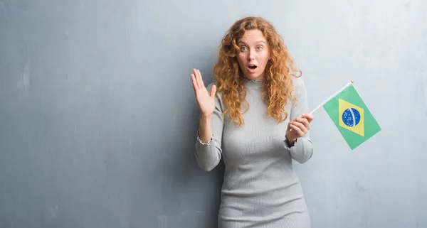 年轻的红头发的女人在灰色的垃圾墙持有巴西国旗非常高兴和兴奋 优胜者表达庆祝胜利尖叫着大微笑和举手 — 图库照片