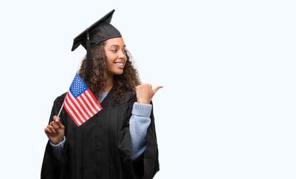 Young hispanic woman wearing graduation uniform holding flag of United States pointing and showing with thumb up to the side with happy face smiling