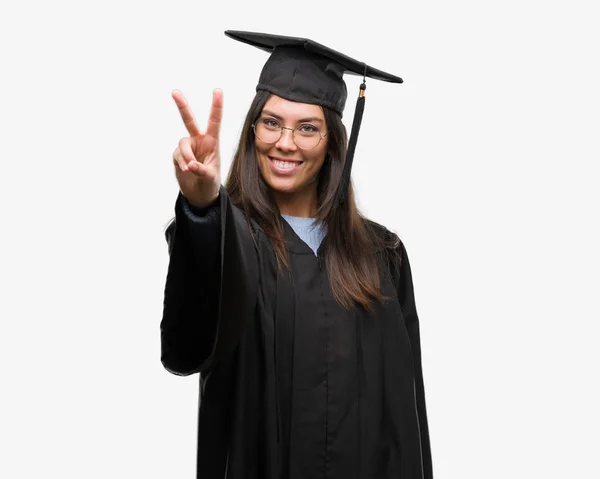 年轻的西班牙裔妇女穿着毕业帽和制服微笑着看着相机显示手指做胜利标志 第二个 — 图库照片
