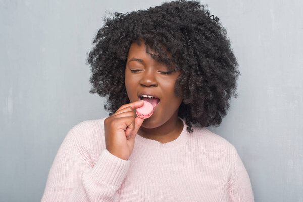 Young african american woman over grey grunge wall eating pink macaron with a confident expression on smart face thinking serious