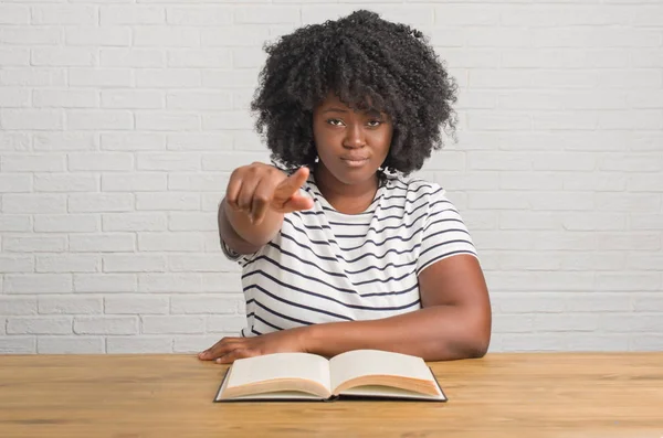 Young african american woman sitting on the table reading a book pointing with finger to the camera and to you, hand sign, positive and confident gesture from the front