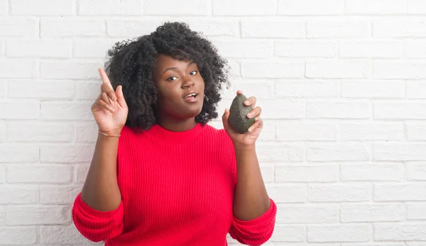 年轻的非洲裔美国人妇女在白色砖墙吃鳄梨惊奇与想法或问题指向手指与愉快的面孔 — 图库照片