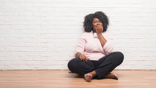 Stock image Young african american woman sitting on the floor wearing headphones looking stressed and nervous with hands on mouth biting nails. Anxiety problem.