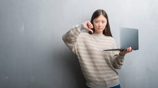 年轻的中国妇女在灰色墙壁使用计算机膝上型电脑与愤怒的面孔 消极标志显示不喜欢与拇指下来 拒绝概念 — 图库照片