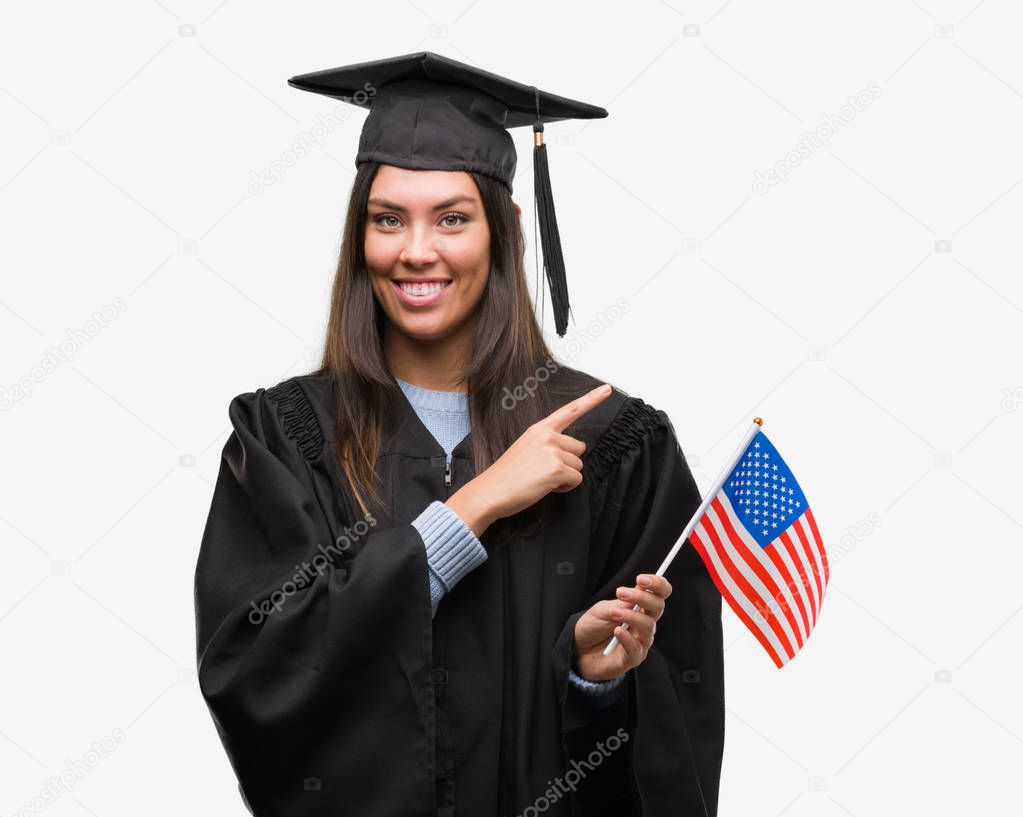 Young hispanic woman wearing graduated uniform holding flag of america very happy pointing with hand and finger to the side