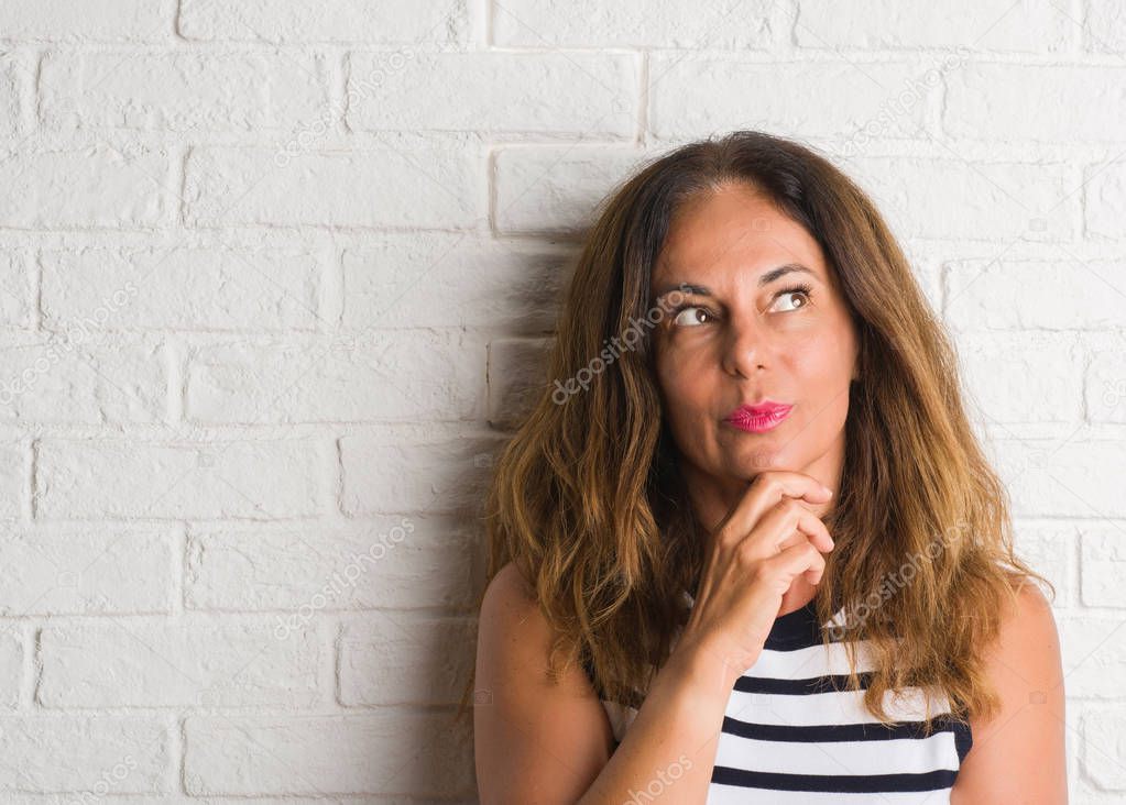 Middle age hispanic woman over white brick wall serious face thinking about question, very confused idea