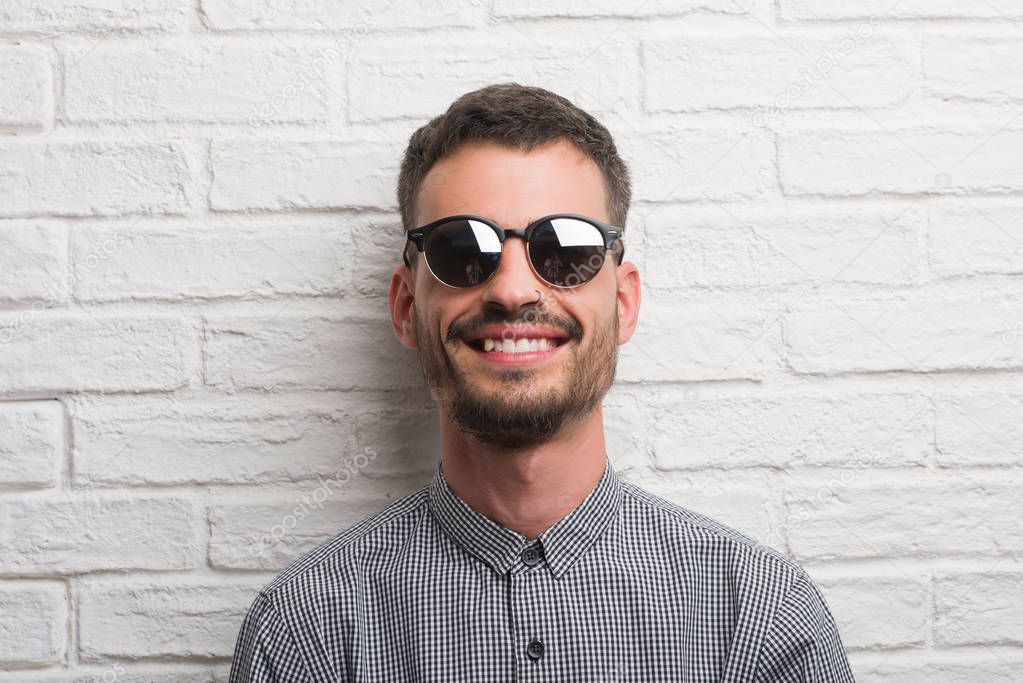 Young adult man wearing sunglasses standing over white brick wall with a happy face standing and smiling with a confident smile showing teeth