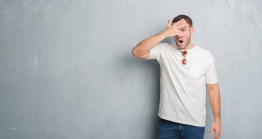 Mahcup ifadeyle parmaklarımın arasından görünen genç beyaz adam yüz ve gözler el ile kapsayan şok bakmak güneş gözlüğü takıyor gri grunge duvarın üzerinden.