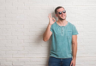 Parmak, mükemmel sembolü ile ok işareti yaparken güneş gözlüğü takmış beyaz tuğla duvarın üzerinden ayakta beyaz delikanlı