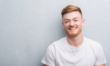 Genç Kızıl saçlı adam ayakta ve diş gösterilen kendine güvenen bir gülümsemeyle gülümseyerek mutlu bir yüz ile gri grunge duvarın üzerinden