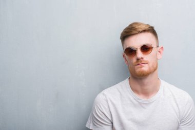 Young redhead man over grey grunge wall wearing retro sunglasses with a confident expression on smart face thinking serious clipart