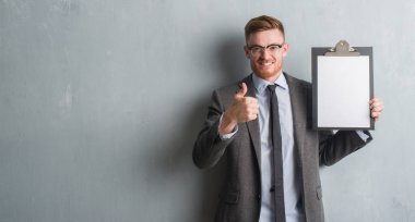 Genç redhead iş adamı ok işareti, başparmak ile parmak, mükemmel işareti yapıyor kocaman bir gülümseme ile pano mutlu tutarak gri grunge duvarın üzerinden