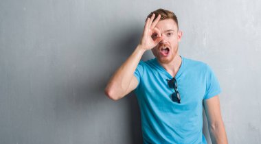 Sürpriz yüz, göz parmaklarının arasından seyir gri grunge duvar ok jest yapıyor gündelik kıyafeti giymiş genç Kızıl saçlı adam şok. İmansız ifade.