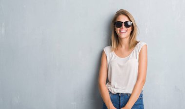 Ayakta ve diş gösterilen kendine güvenen bir gülümsemeyle gülümseyerek mutlu bir yüz ile moda güneş gözlüğü takıyor grunge gri duvar üzerinde duran güzel genç kadın