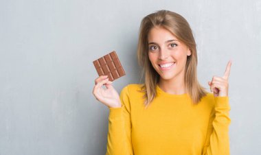 Grunge güzel genç kadın çikolatayı bir fikirle sürpriz yemek duvar gri veya gülen yüz, bir numara ile işaret parmağı soru