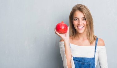 Güzel genç kadın ayakta ve diş gösterilen kendine güvenen bir gülümsemeyle gülümseyerek mutlu bir yüz ile nar tutan grunge gri duvarın üzerinden