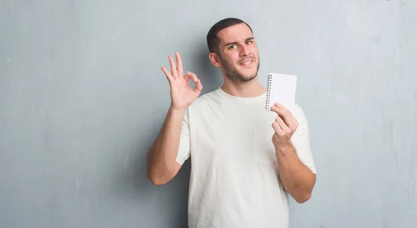 年轻的白种人在灰色垃圾墙显示笔记本做 标志与手指 优秀的符号 — 图库照片