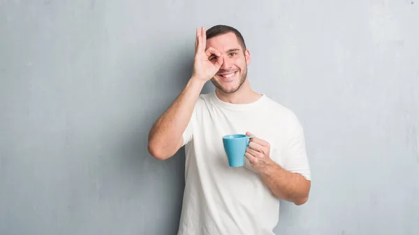 年轻的白种人在灰色垃圾墙壁喝一杯咖啡与愉快的面孔微笑做 标志用手在眼睛看通过手指 — 图库照片