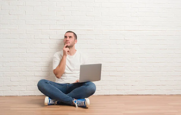 年轻的白种人坐在白色砖墙上使用电脑笔记本电脑严重的面孔思考问题 非常困惑的想法 — 图库照片