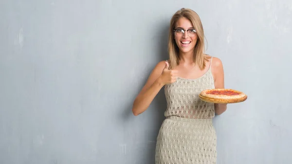 グランジに美しい若い女性灰色壁笑顔の サイン 優秀な兆候を親指をやって幸せなペパロニのピザを食べて — ストック写真