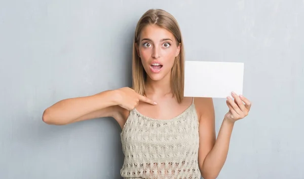 Beautiful young woman over grunge grey wall holding blank card with surprise face pointing finger to himself
