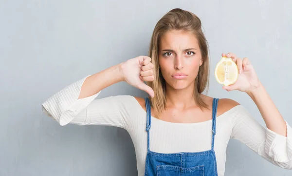 美丽的年轻女子在破烂的灰色墙壁拿着一个柠檬与愤怒的面孔 消极标志显示不喜欢与拇指下来 拒绝概念 — 图库照片