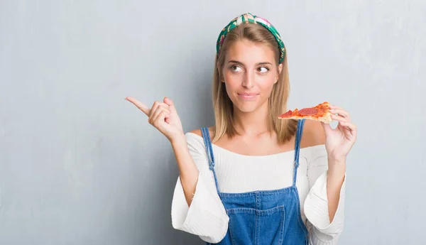グランジ灰色壁側の手と指で指す非常に幸せなペパロニのピザのスライスを食べて美しい若い女性 — ストック写真