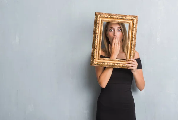 Beautiful young woman over grunge grey wall holding vintage frame cover mouth with hand shocked with shame for mistake, expression of fear, scared in silence, secret concept