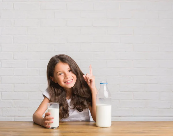 幸せそうな顔でのアイデアや質問ポインティング指に驚いてミルクのガラスを飲むテーブルに座って若いヒスパニックの子供がナンバーワンします — ストック写真