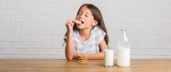 牛乳を飲むと考えて深刻なスマートの顔に自信を持って式にチョコレート クッキーを食べるテーブルに座って若いヒスパニックの子供 — ストック写真