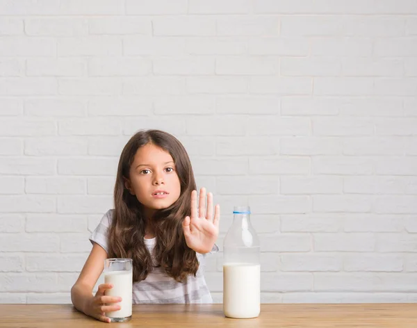 ミルクのガラスを飲むテーブルに座って若いヒスパニックの子供は深刻なと自信を持って式 防衛ジェスチャーと一時停止の標識をやって手を開いてください — ストック写真
