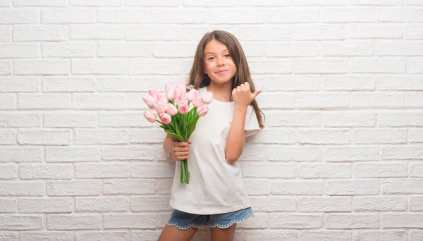 指していると幸せそうな顔を笑顔で側に親指を示す母の日に花を持って白いレンガ壁にヒスパニック系の子供 — ストック写真