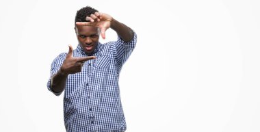 Genç Afro-Amerikan adam elleriyle yapım çerçeve ve parmakları ile mutlu yüz gülümseyen mavi tişört giyiyor. Yaratıcılık ve Fotoğrafçılık kavramı.