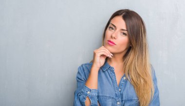 Grunge gri duvar çene düşünme hakkında soru, dalgın ifade üzerine el ile denim kıyafeti giymiş genç yetişkin kadın. Düşünceli yüzüyle gülümsüyor. Şüphe kavramı.